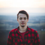 Porträt eines dreiundzwanzigjährigen Mannes in einem schwarz-rot karierten Hemd, der seinen Freund mit seinem echten Lächeln ohne jede Verstellung anlächelt. Die authentische Stimmung eines jungen Mannes auf einer unentschlossenen Reise